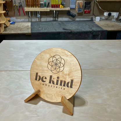 Wooden sign with 'be kind' Industries logo on a workshop table